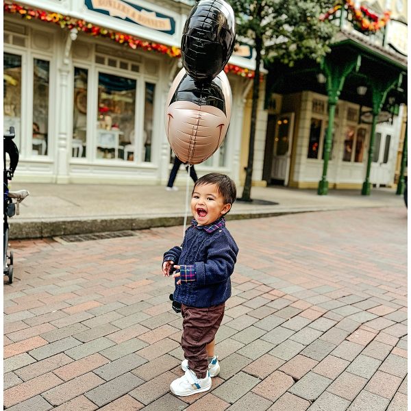 A happy toddler holding a Mickey Mouse balloon, laughing and playing on Main Street at Disneyland Paris. The child is dressed warmly in a navy sweater and brown pants, with the iconic Disney storefronts and festive decorations visible in the background. The photo captures the magical experience and joy of visiting Disneyland Paris with young children.